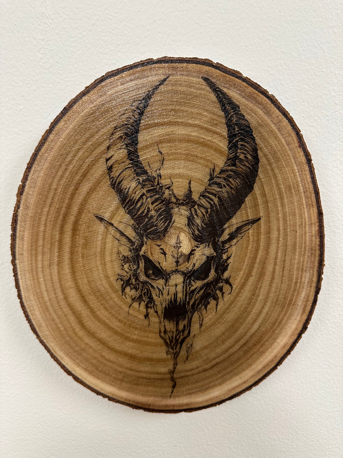 Wooden plaque with a black ink drawing of a goat skull and horns on a white background