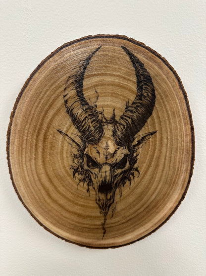 Wooden plaque with a black ink drawing of a goat skull and horns on a white background