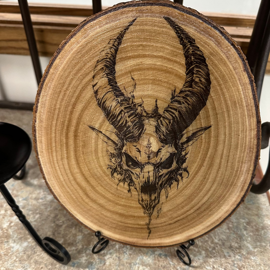 Close Up - Front View on a stand
Round-Wooden piece with a black ink drawing of a goat skull with horns on a metal stand.